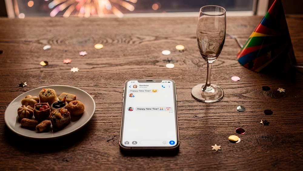 new years eve texting call playbook — top-down view of a phone with a short New Year’s Eve text draft next to a glass and small fireworks outside the window in the background